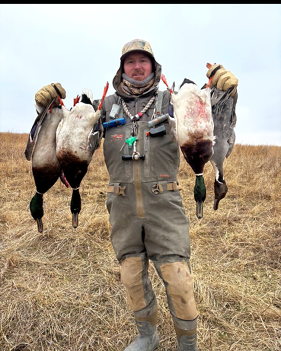 Joey Benton in waders holding a strap of drake mallards and a pintail